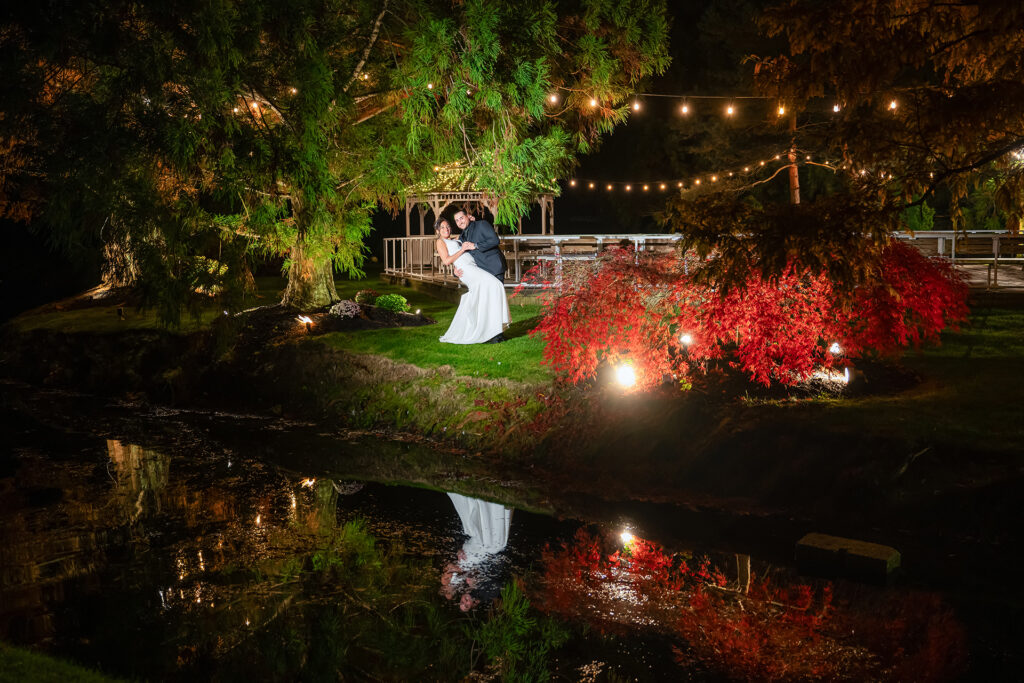 Bride and groom posing under string lights by a pond with reflections at Flowerfield Celebrations in St James NY during a nighttime wedding portrait