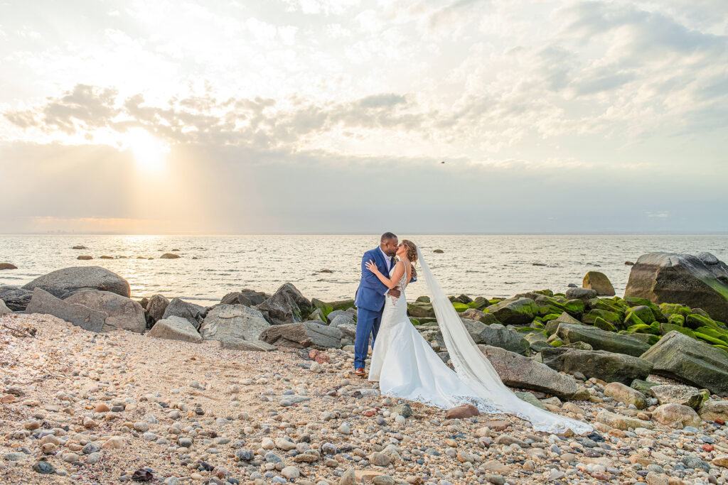 Bride and groom kissing on a rocky beach at sunset at Soundview Caterers in Bayville NY with ocean waves and dramatic sky in the background