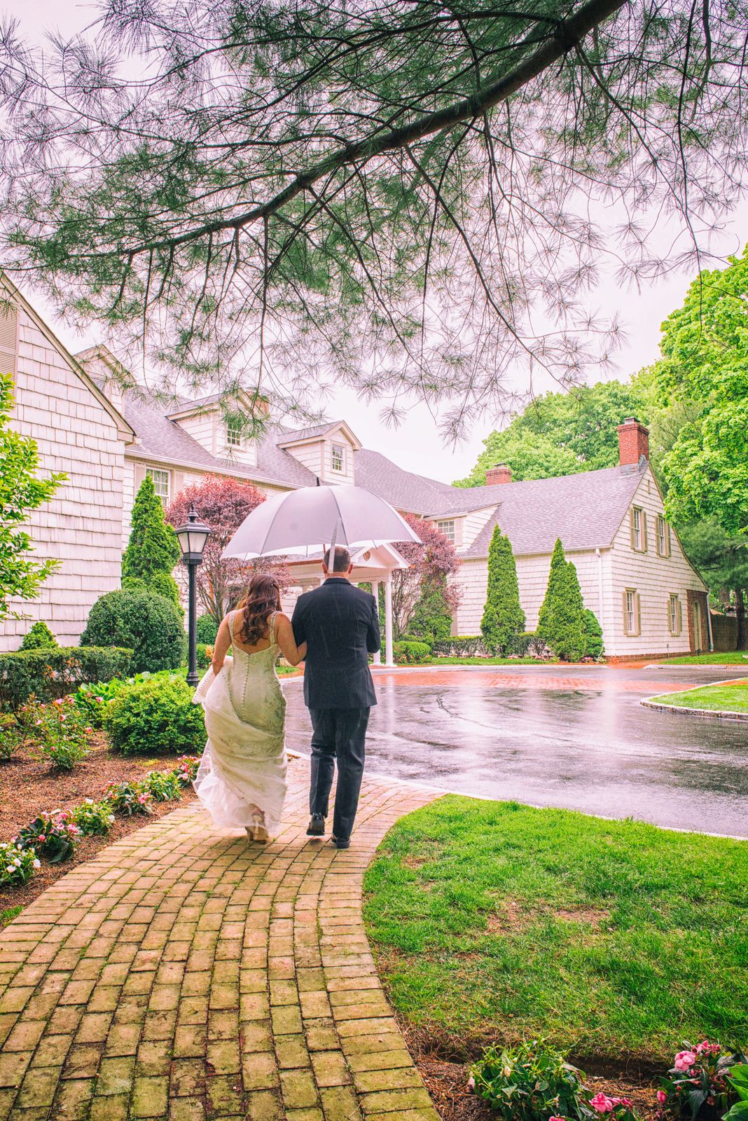 Bride and groom walk under an umbrella along the garden path at The Royalton Mansion