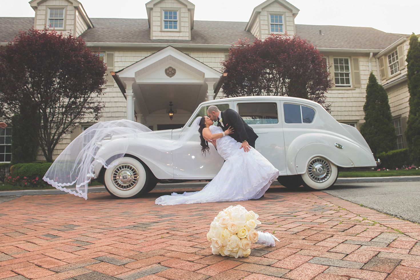 Bride and groom kiss beside a vintage white car at The Royalton Mansion