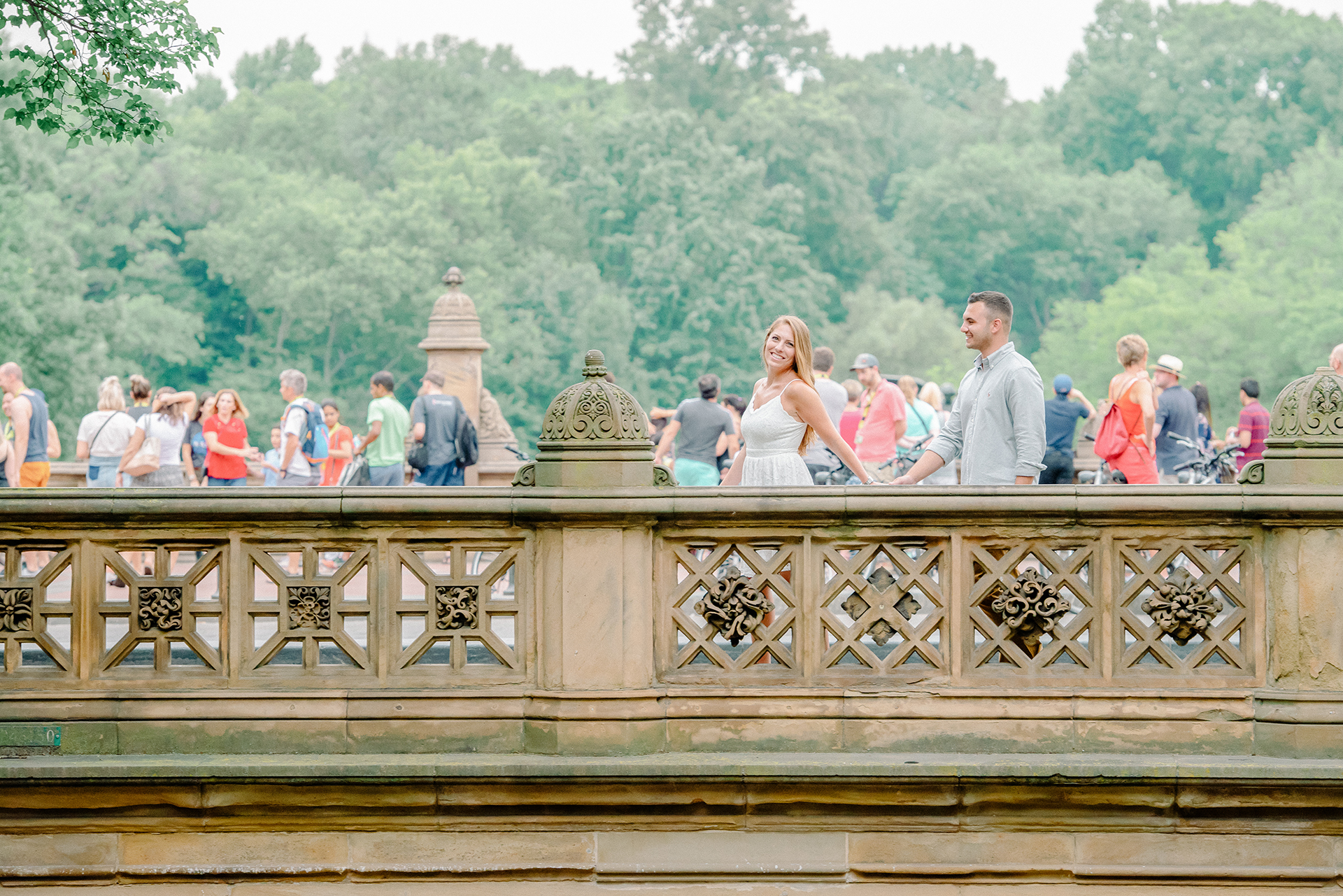Couple smiling and holding hands near the Bow Bridge area of Central Park with visitors in the background.