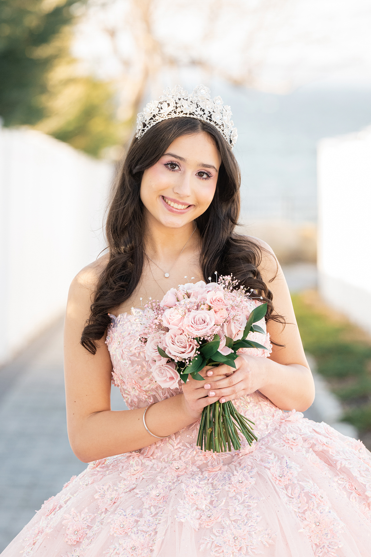 Quinceañera sonriente sosteniendo un ramo de rosas rosadas, con una tiara brillante y un vestido rosa pálido.