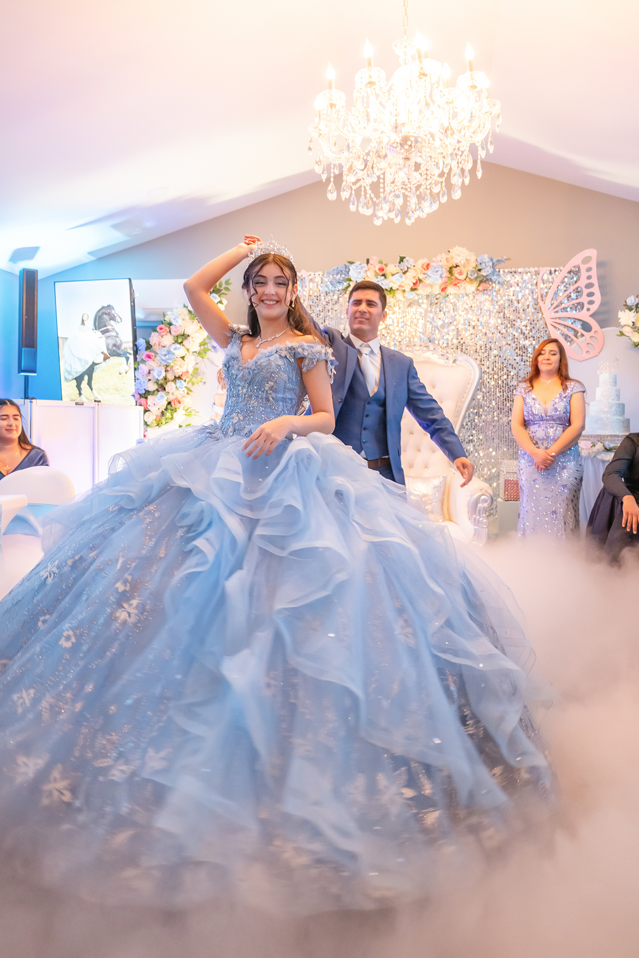 Quinceañera haciendo su gran entrada con un vestido azul de gala, rodeada de familia y efectos de hielo seco.