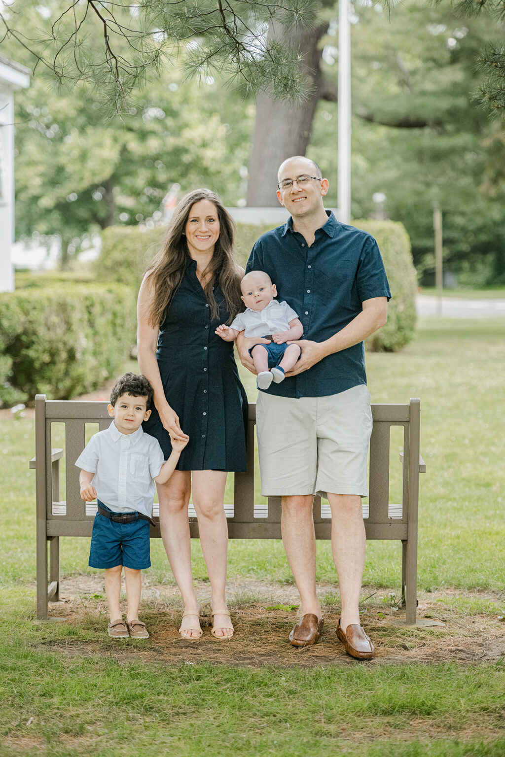 Family of four standing near a park bench during an outdoor portrait session surrounded by greenery.