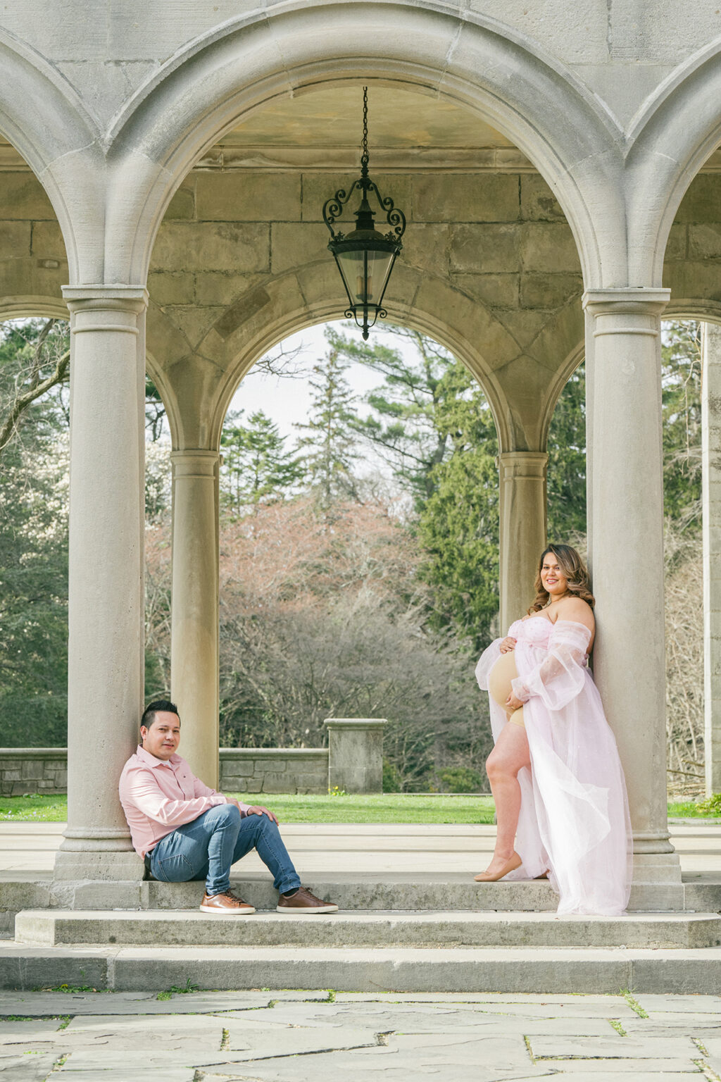 Expectant mother in a flowing pink gown posing with her partner beneath large stone arches.