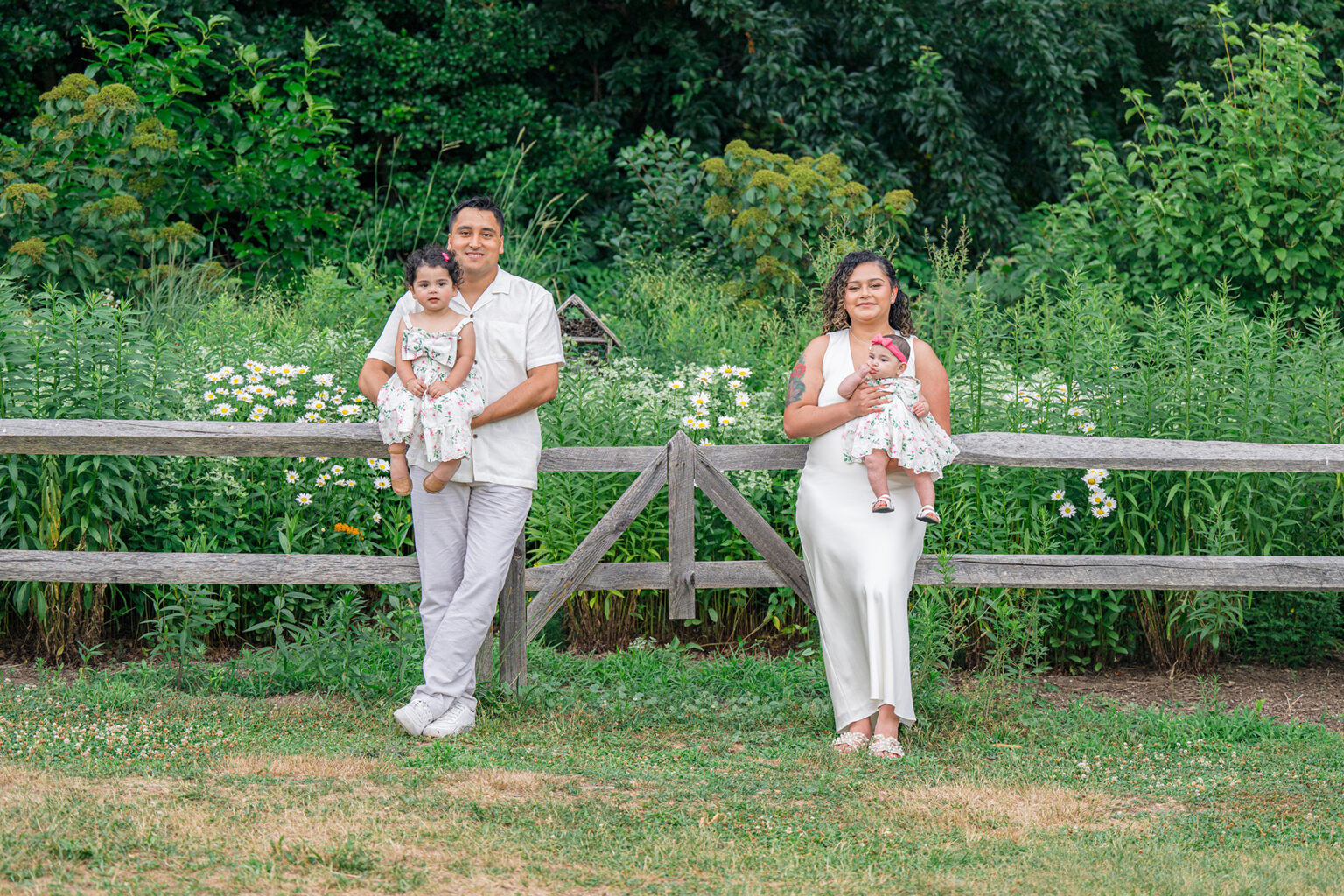 Parents holding their two young daughters in matching floral dresses beside a wooden garden fence.