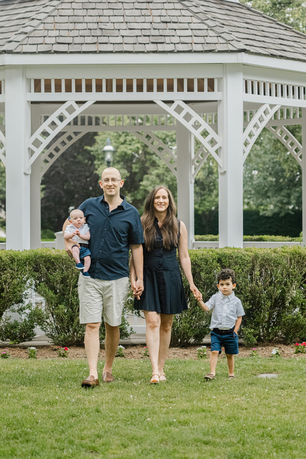 Parents walking hand in hand with their two children in front of a white garden gazebo.