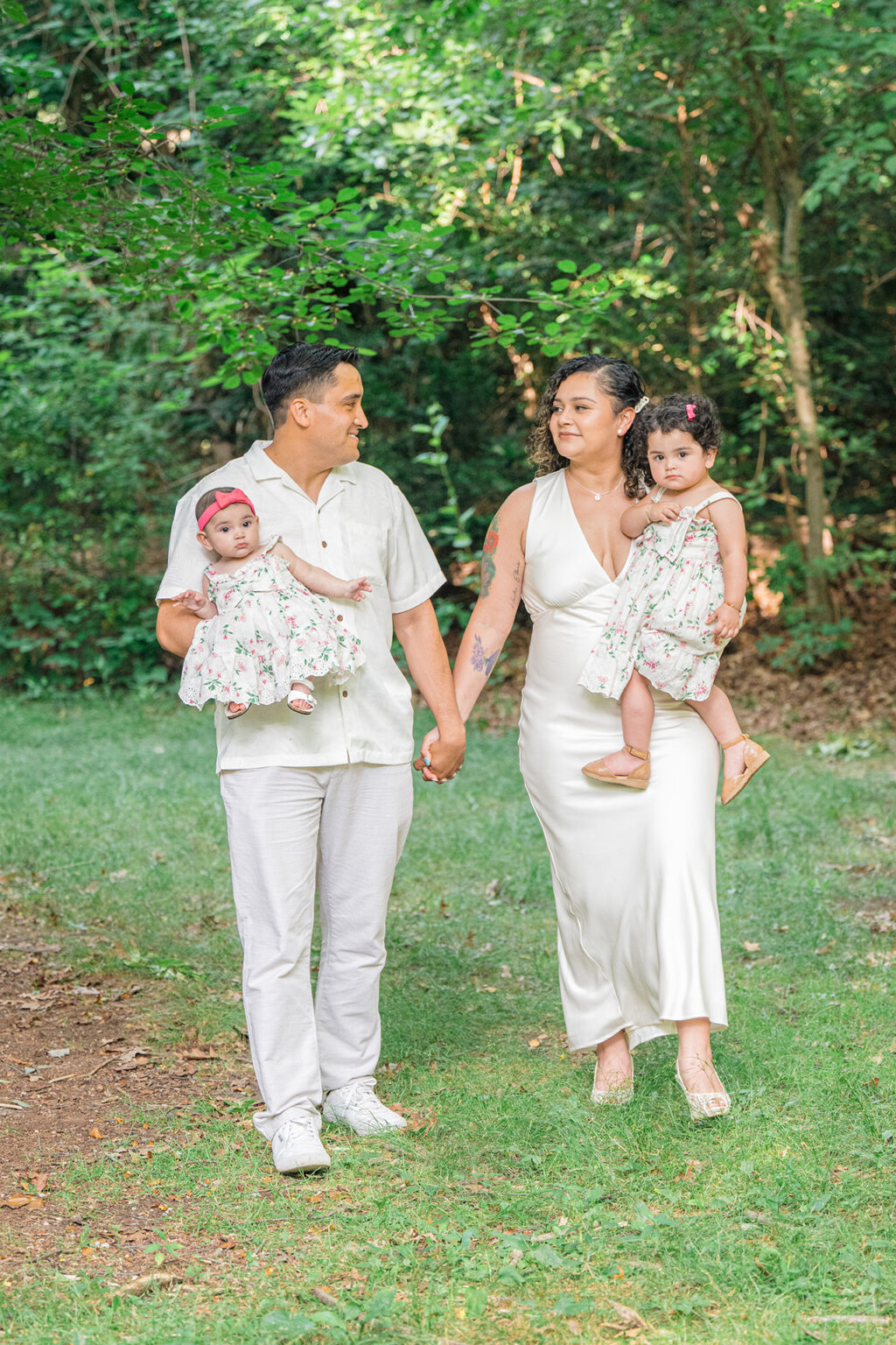 Parents carrying their two young daughters while walking together through a wooded park.