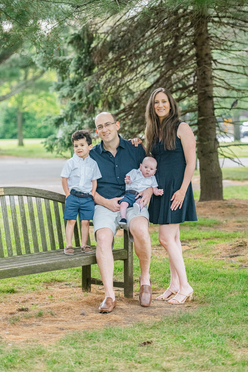 Family portrait of parents with two young sons beside a wooden bench in a green Long Island park.