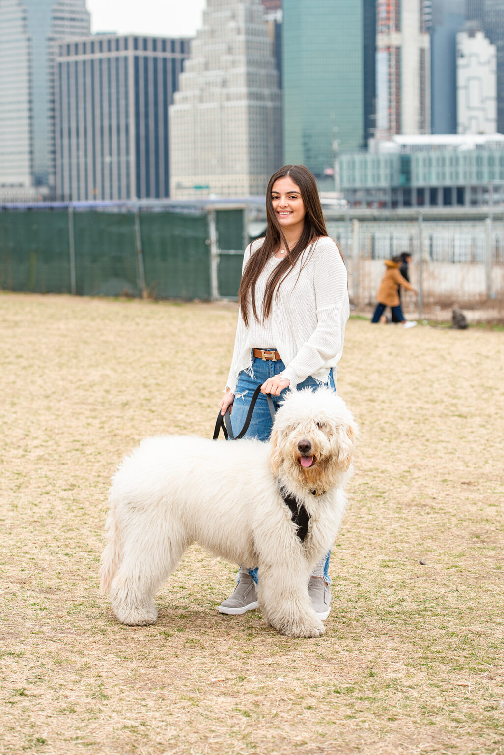 Young woman posing with a large white dog near the Brooklyn waterfront and New York City skyline.