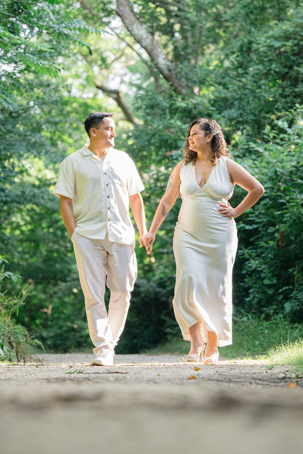 Couple holding hands and walking along a sunlit wooded path during an outdoor portrait session.