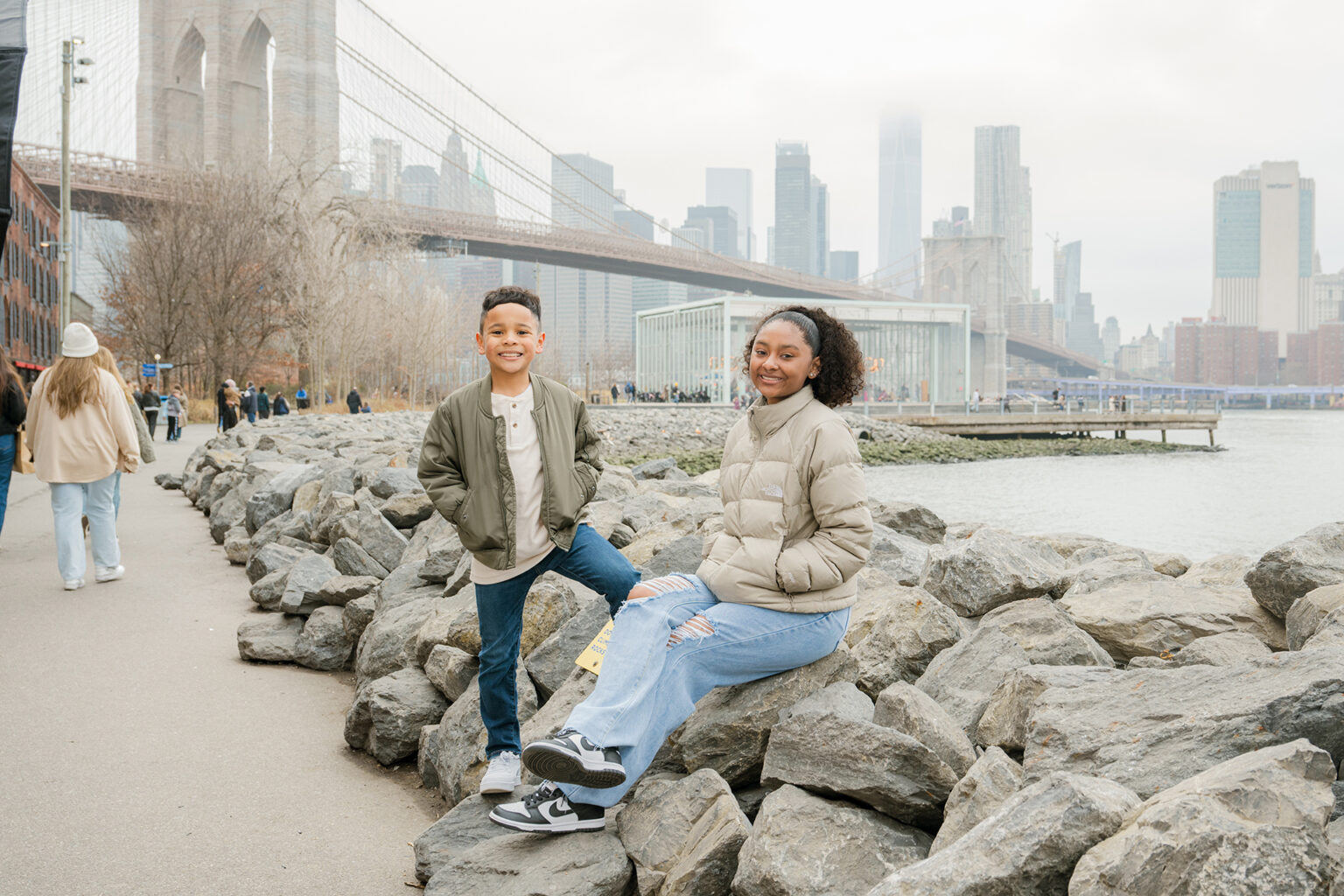 Brother and sister posing on waterfront rocks with the Brooklyn Bridge and Manhattan skyline behind them.